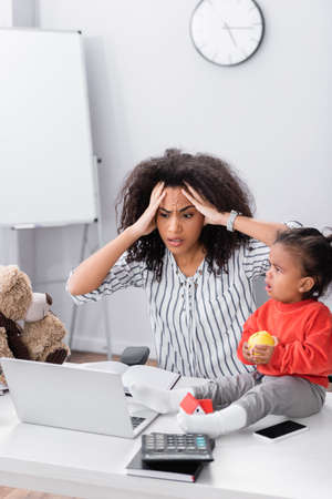 stressed african american freelancer near toddler daughter sitting on desk with appleの写真素材