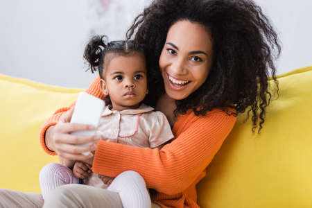 happy african american mother holding smartphone and taking selfie with toddler daughterの写真素材