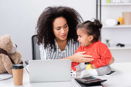 curly african american freelancer supporting toddler daughter sitting on desk near laptopの写真素材