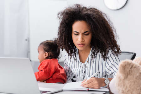 stressed african american mother holding in arms crying toddler daughter while working from homeの写真素材