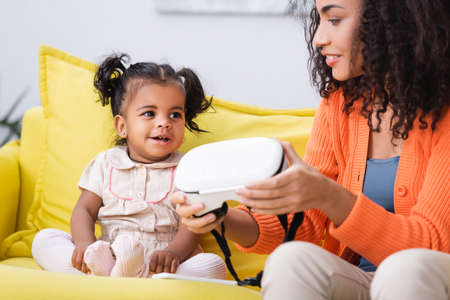 happy african american mother holding vr headset near daughter in living roomの写真素材