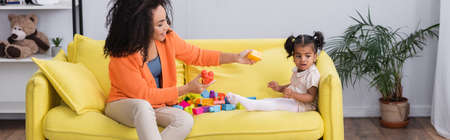 happy african american mother playing building blocks with toddler kid on sofa, banner