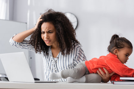 stressed african american mother adjusting hair and looking at laptop near toddler kid crawling on deskの写真素材