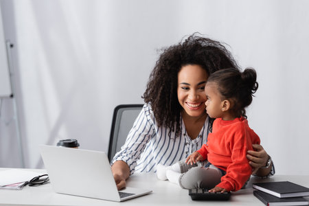 smiling african american mother looking at toddler daughter sitting near laptop on deskの写真素材