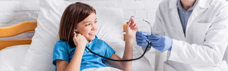 doctor showing stethoscope to cheerful girl in hospital bed, bannerの写真素材