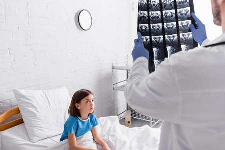 doctor holding x-ray near child sitting on bed in hospital, blurred foreground