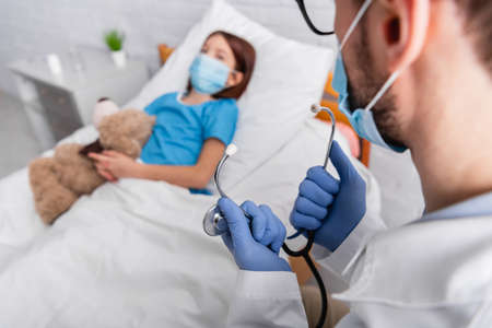 doctor holding stethoscope near sick child in medical mask lying in bed with teddy bear on blurred...