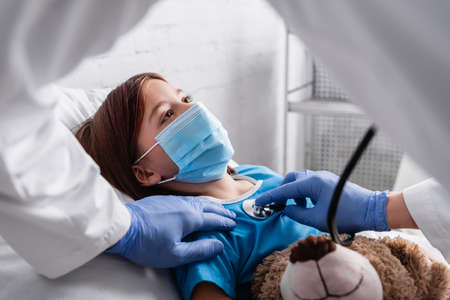 diseased girl in medical mask lying in bed with teddy bear while pediatrician examining her with stethoscope, blurred foregroundの写真素材