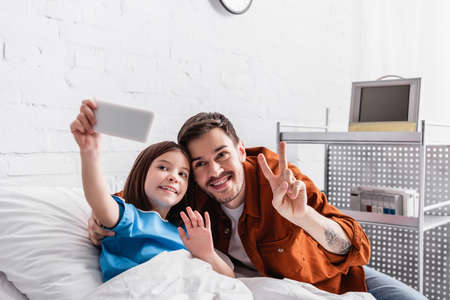 happy man showing victory gesture near daughter waving hand while taking selfie on smartphone,...