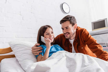 happy man hugging smiling daughter talking on smartphone in hospital bed