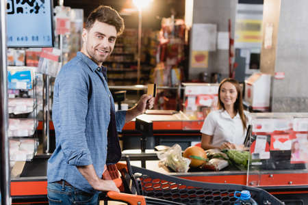 Smiling man holding credit card near shopping cart and supermarket checkout on blurred background