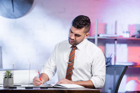 concentrated architect writing in notebook near models of wind generators