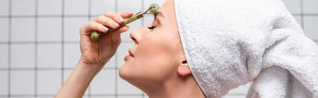 side view of woman with towel on head using jade roller in bathroom, bannerの写真素材