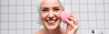 cheerful woman in towel on head using cleansing silicone brush in bathroom, bannerの写真素材
