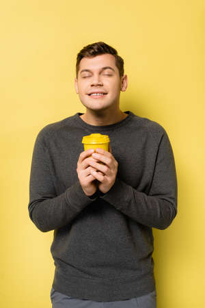 Happy man with closed eyes holding coffee to go on yellow backgroundの写真素材