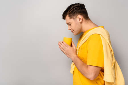 Side view of young man smelling drink in yellow cup on gray background