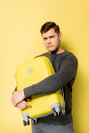 Displeased man hugging suitcase on yellow backgroundの写真素材