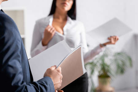 partial view of businessman with paper folder near seductive businesswoman showing bust in office,...