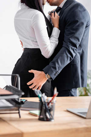 partial view of businessman embracing and kissing businesswoman on blurred foreground in officeの写真素材
