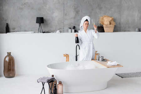 Young woman in bathrobe holding bottle and glass of champagne in bathroom