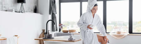 Woman in bathrobe applying cosmetic cream on leg in bathroom, banner