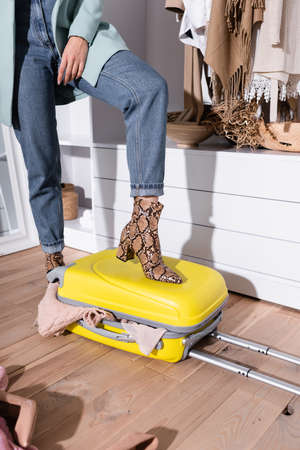 Cropped view of woman putting feet on suitcase with clothes in wardrobe