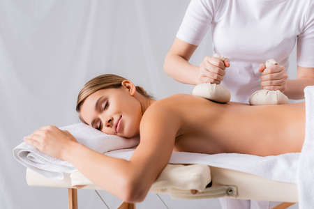woman massaging young pleased client with herbal bags in spa salonの写真素材