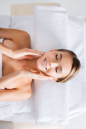 top view of happy woman touching cheeks and lying on massage table in spa salonの写真素材
