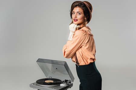 stylish woman with hands near face looking at camera near record player isolated on grayの写真素材