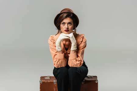 pretty, trendy woman sitting on vintage suitcase while looking at camera on gray