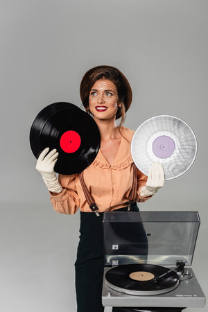 happy woman in vintage clothes holding vinyl discs near record player isolated on gray