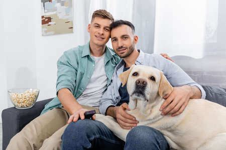 cheerful same sex couple smiling near bowl with popcorn and labradorの写真素材