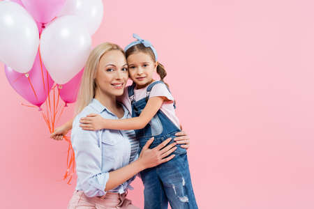 happy kid holding balloons and hugging cheerful mother isolated on pink