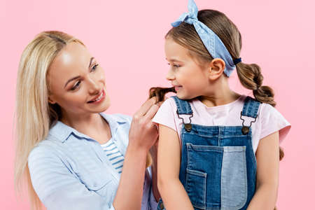 happy mother braiding hair of daughter isolated on pink