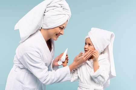 happy mother in bathrobe holding tube and applying cosmetic cream on face of daughter isolated on blueの写真素材