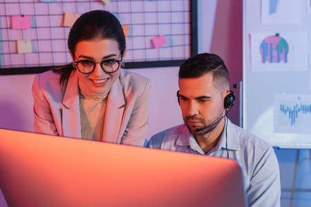 operator in headset looking at computer monitor near smiling businesswomanの写真素材