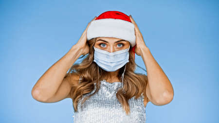 woman with wavy hair in medical mask wearing santa hat isolated on blueの写真素材