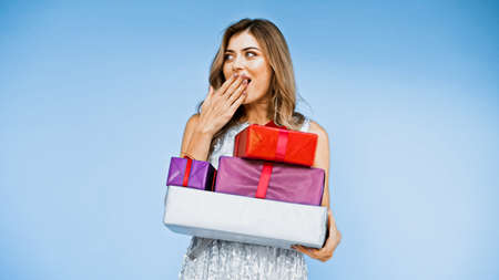 happy woman covering mouth while holding wrapped presents isolated on blueの写真素材