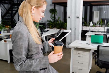 Side view of businesswoman with coffee to go holding digital tablet in office