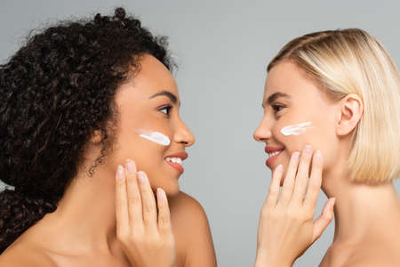 Young multicultural women with naked shoulders and cosmetic cream on cheeks looking at each other...