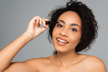 Smiling african american woman with naked shoulders applying mascara isolated on grayの写真素材