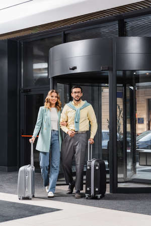 Young interracial couple with baggage looking at camera near hotel