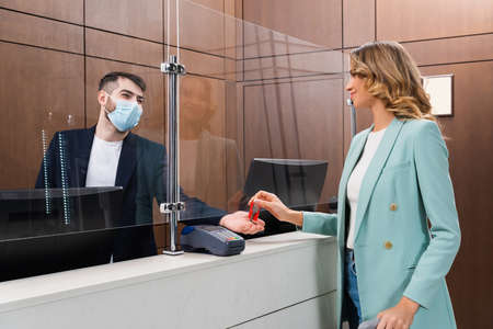 Hotel manager in medical mask giving key to young woman in lobby