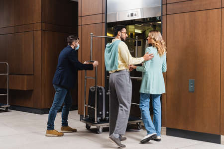 Interracial couple standing near hotel porter in medical mask and elevatorの写真素材