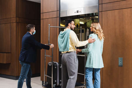 Interracial couple smiling near porter in medical mask and elevator in hotel lobby