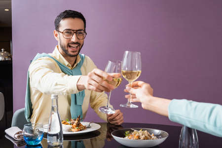 Cheerful arabian man clinking wine with girlfriend near dinner in hotel room