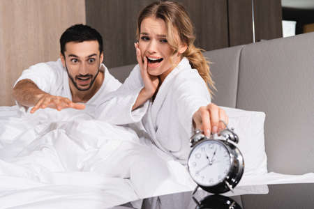 Worried woman and middle eastern man looking at alarm clock on blurred foreground in hotel