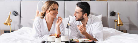 Smiling woman looking at middle eastern boyfriend near breakfast on bed in hotel room, banner