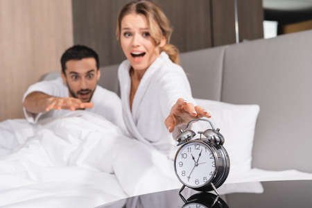 Alarm clock near excited woman and muslim man on hotel bed on blurred backgroundの写真素材