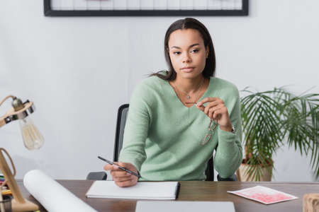 pensive african american interior designer holding pencil and eyeglasses while sitting near...